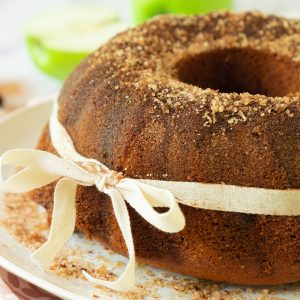 A bundt cake with a cinnamon sugar topping, wrapped with a cream-colored ribbon tied in a bow, sits on a plate.