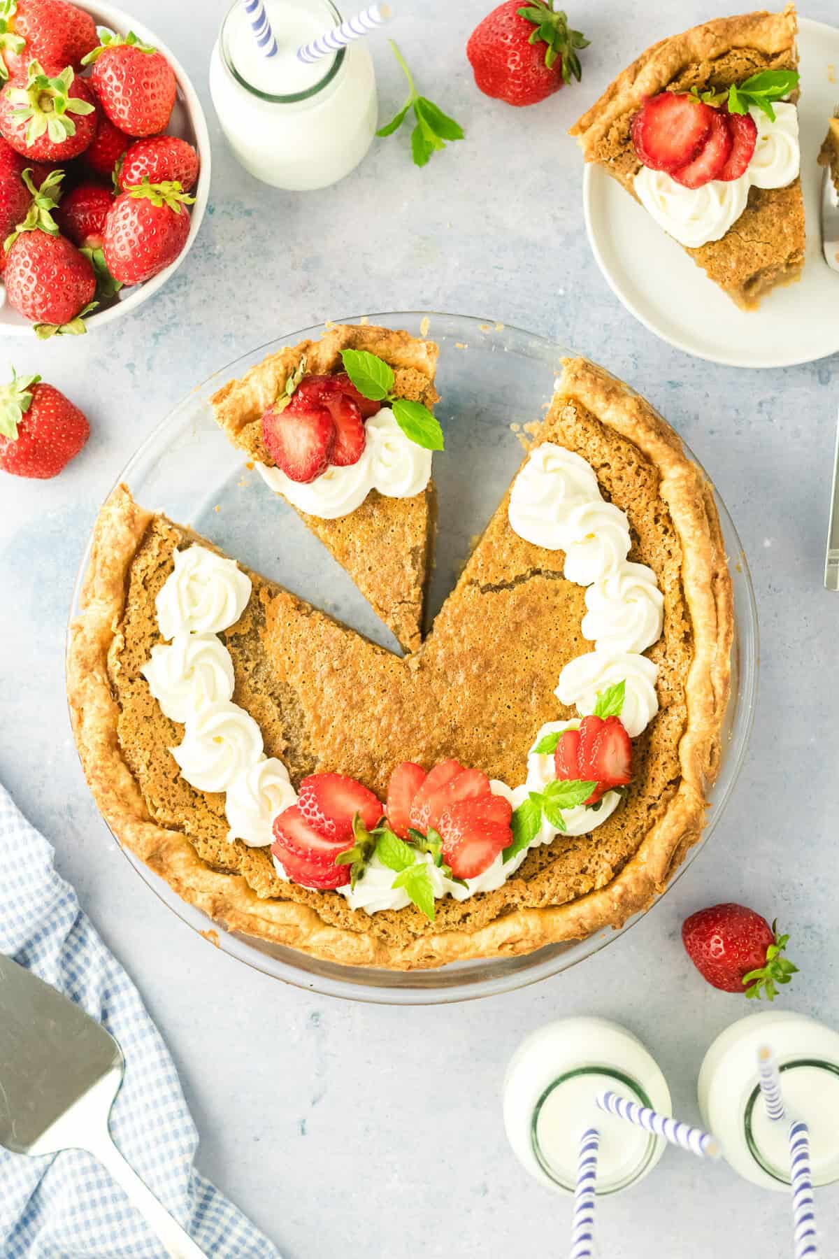 A baked pie with a golden crust sits on a table, topped with whipped cream, sliced strawberries, and mint leaves. One slice is served on a plate; fresh strawberries and bottles of milk with straws are nearby.