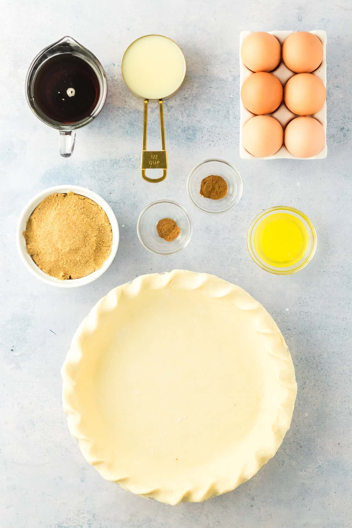 Unbaked pie crust in a dish surrounded by eggs, melted butter, brown sugar, spices in small bowls, sweetened condensed milk, and dark syrup in a measuring jug, all on a light surface.