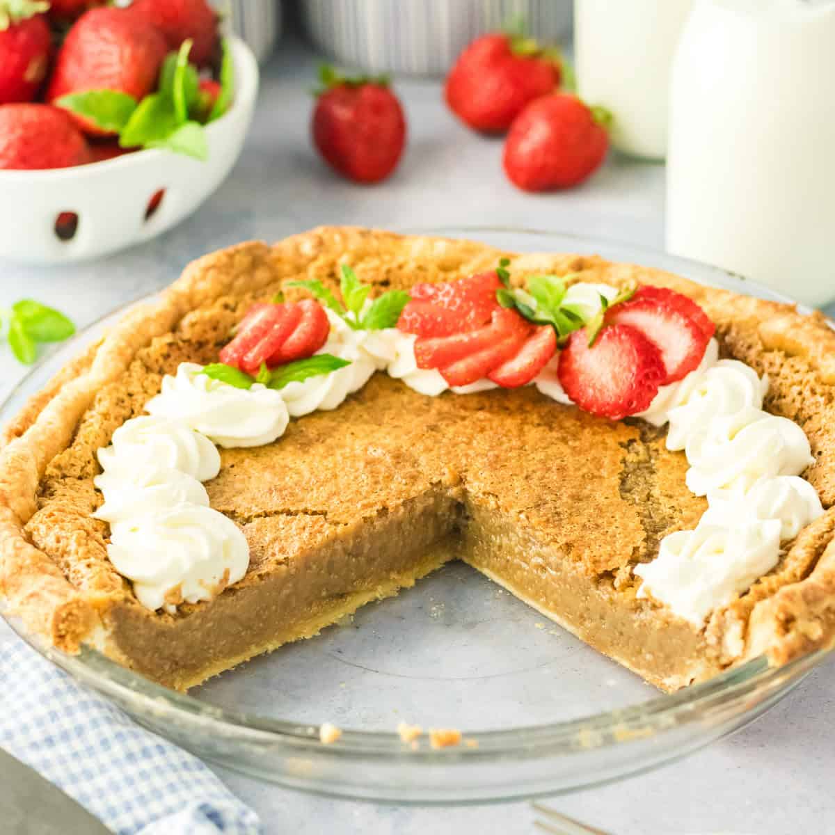 A pie in a glass dish with a golden-brown filling, topped with dollops of whipped cream and sliced strawberries. A slice has been removed, revealing the pies soft interior. Fresh strawberries and bottles of milk are in the background.