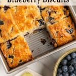 A pan of golden, baked blueberry biscuits with one square partially removed. Fresh blueberries and lemon wedges are nearby. Text on the image reads Old Fashioned Blueberry Biscuits.