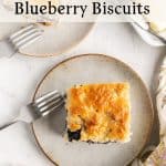 A blueberry biscuit square on a plate with a fork, surrounded by plump blueberries, lemon slices, butter, and another plate with a half-eaten blueberry biscuit. Text reads An Elegant Breakfast: Blueberry Biscuits.