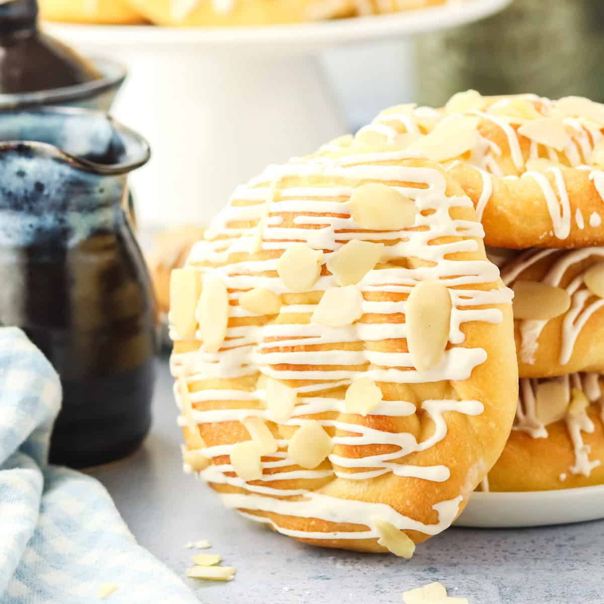 A close-up of round pastries drizzled with white icing and topped with sliced almonds, stacked on a white plate. A blue checkered cloth and a dark ceramic pitcher are nearby.