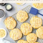 A cooling rack holds several Danish butterhorn pastries drizzled with icing. Nearby are a blue spatula, sliced almonds, a small pitcher of cream, coffee, and a folded blue checked napkin.