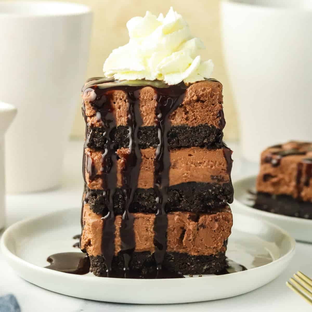 A tall slice of no-bake chocolate cheesecake bars with cookie crust layers, topped with whipped cream and drizzled with chocolate sauce, sits on a white plate.
