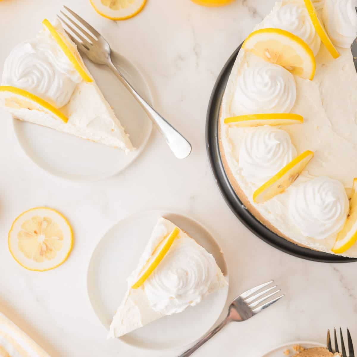 A lemon cheesecake with whipped cream and lemon slices is sliced, with two pieces served on plates, each with a fork nearby, on a white marble surface.