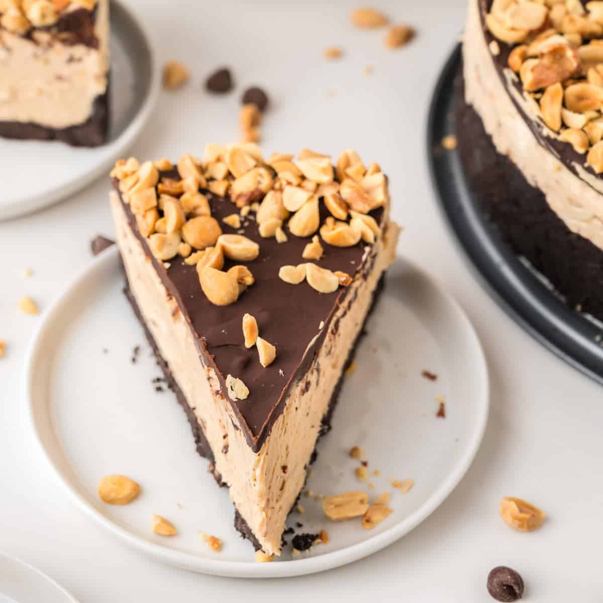 A slice of peanut butter cheesecake topped with chopped peanuts and chocolate sits on a white plate, with more cheesecake and scattered peanuts in the background.