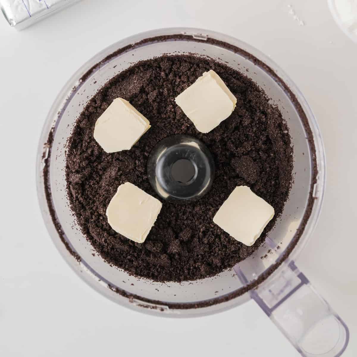 A food processor with crushed chocolate cookies and four cubes of butter on top, ready to be mixed together.