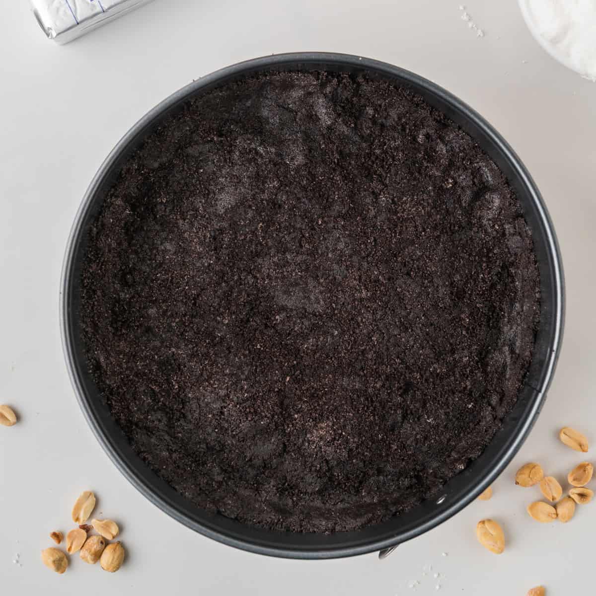 Round springform pan filled with a dark, crumbly Oreo cookie crust. A few peanuts and crumbs are scattered on the white surface beside the pan.