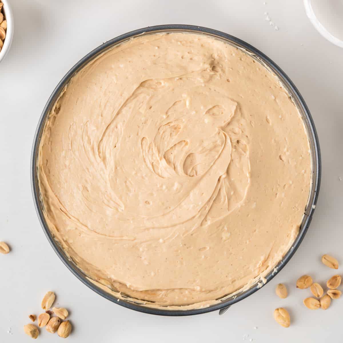 A round pan filled with creamy, light brown batter sits on a white surface, surrounded by scattered peanuts.