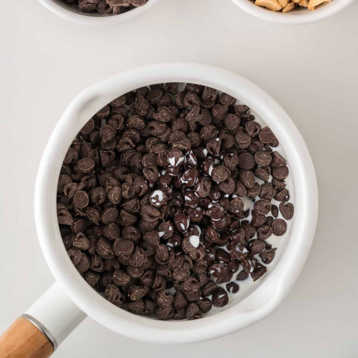 A white saucepan filled with chocolate chips is beginning to melt in the center. Part of a bowl of peanuts and another bowl are visible at the top edge of the image.