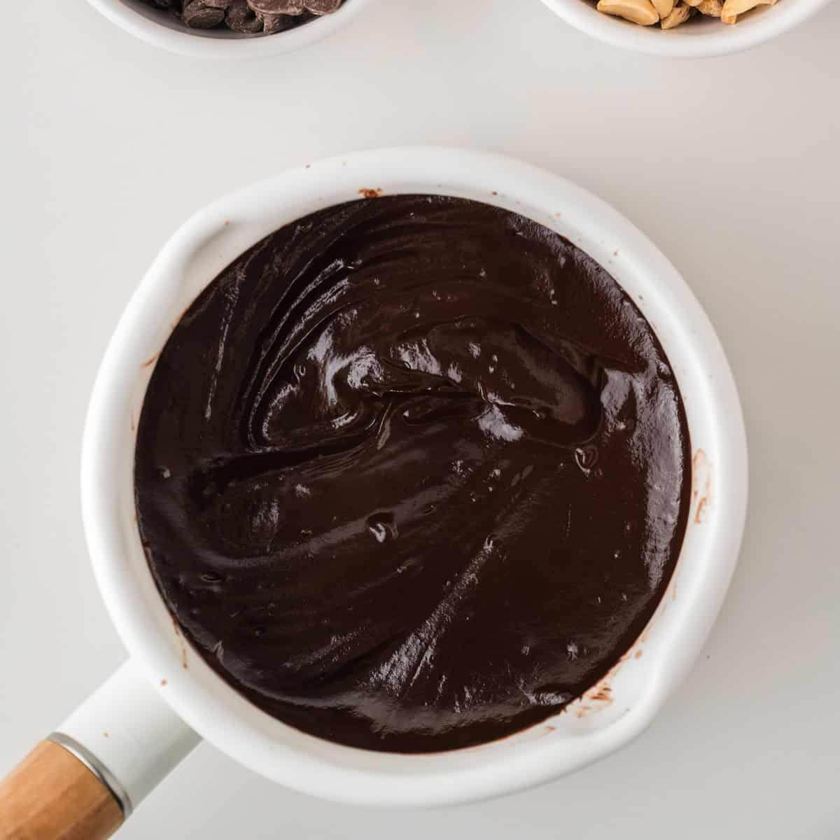 A white saucepan filled with smooth, glossy melted chocolate sits on a white surface, with bowls of chocolate chips and nuts partially visible at the top.