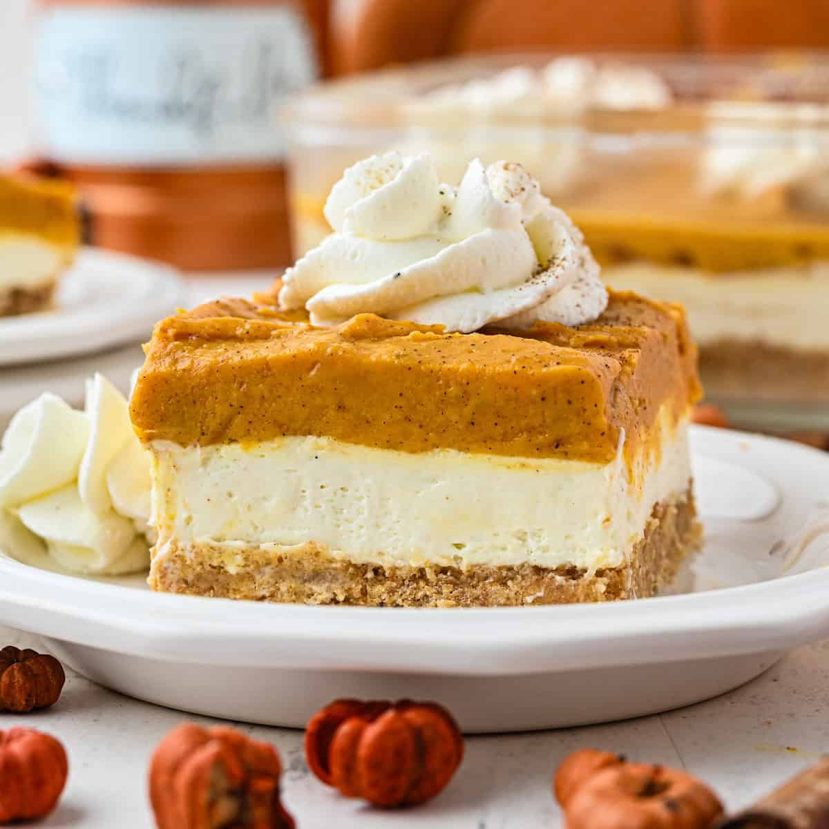 A slice of layered pumpkin dessert with whipped cream on top sits on a white plate, surrounded by small decorative pumpkins. Another dessert and whipped cream are visible in the background.