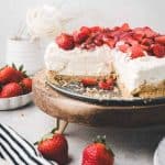 A creamy cheesecake topped with fresh sliced strawberries sits on a wooden cake stand. A slice is missing, revealing the graham cracker crust. Whole strawberries and a striped towel rest nearby.