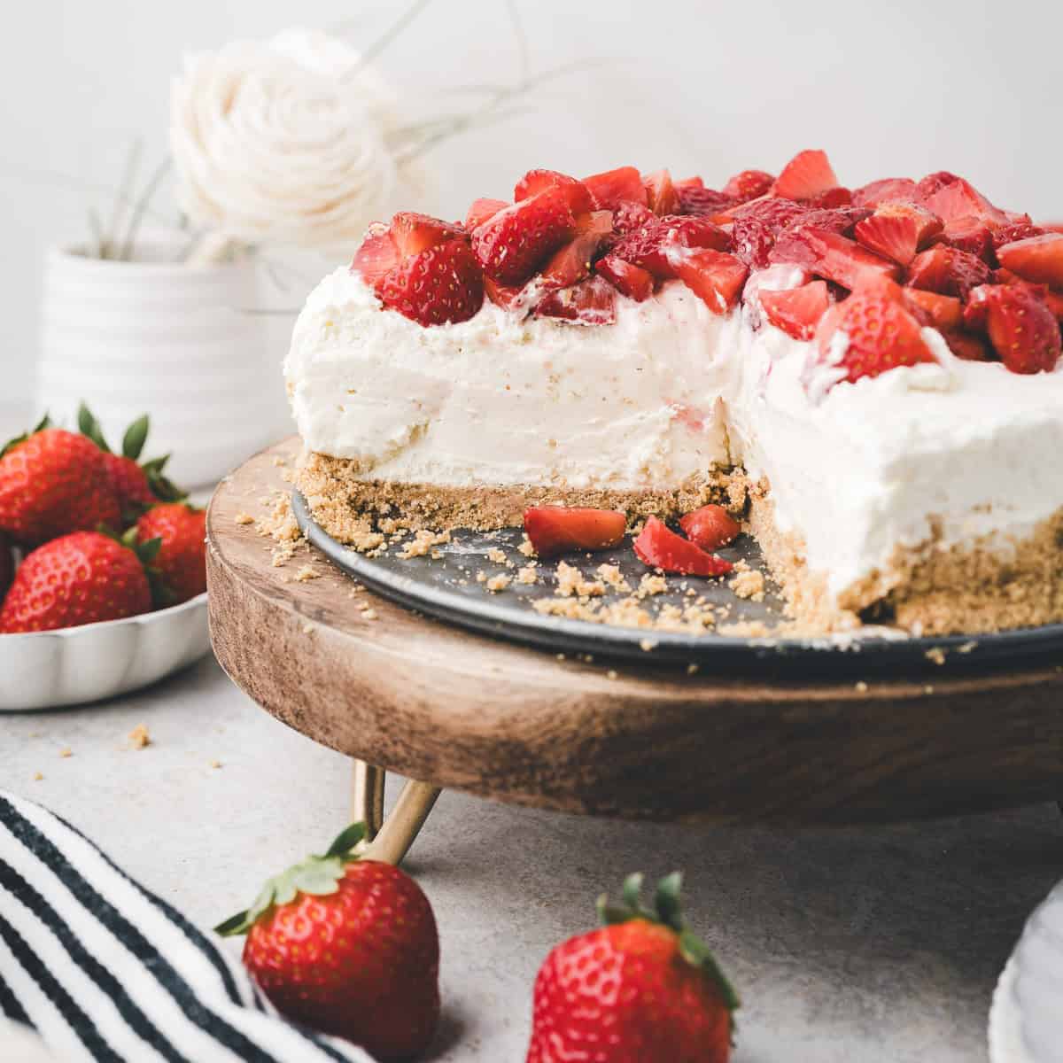 A no-bake cheesecake topped with fresh strawberries sits on a wooden cake stand. A slice is missing, showing the creamy inside. Whole strawberries and crumbs are on the table nearby.
