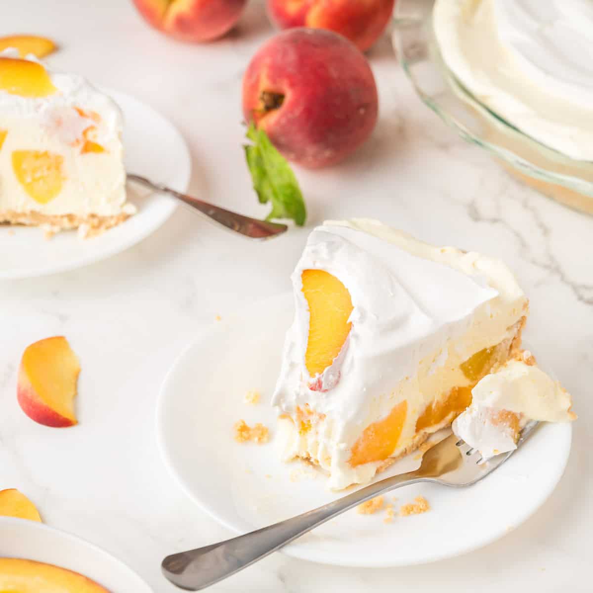 A slice of creamy peach pie with whipped topping on a white plate, a fork, fresh peaches, and another pie slice in the background on a marble surface.