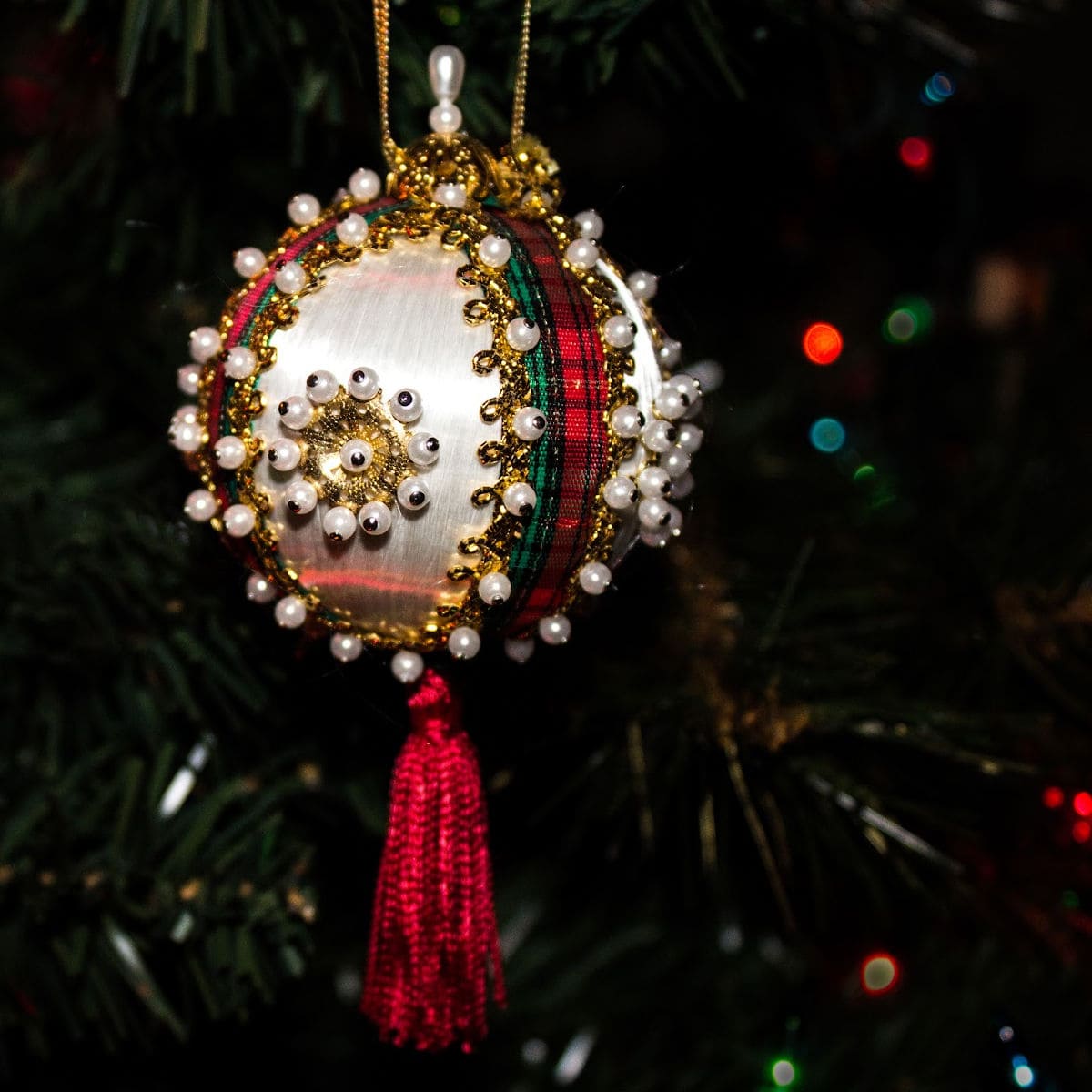 A close-up of an ornate push-pin Christmas ornament decorated with pearls, gold beads, and red ribbon, hanging from a tree branch with blurred colorful lights in the background.