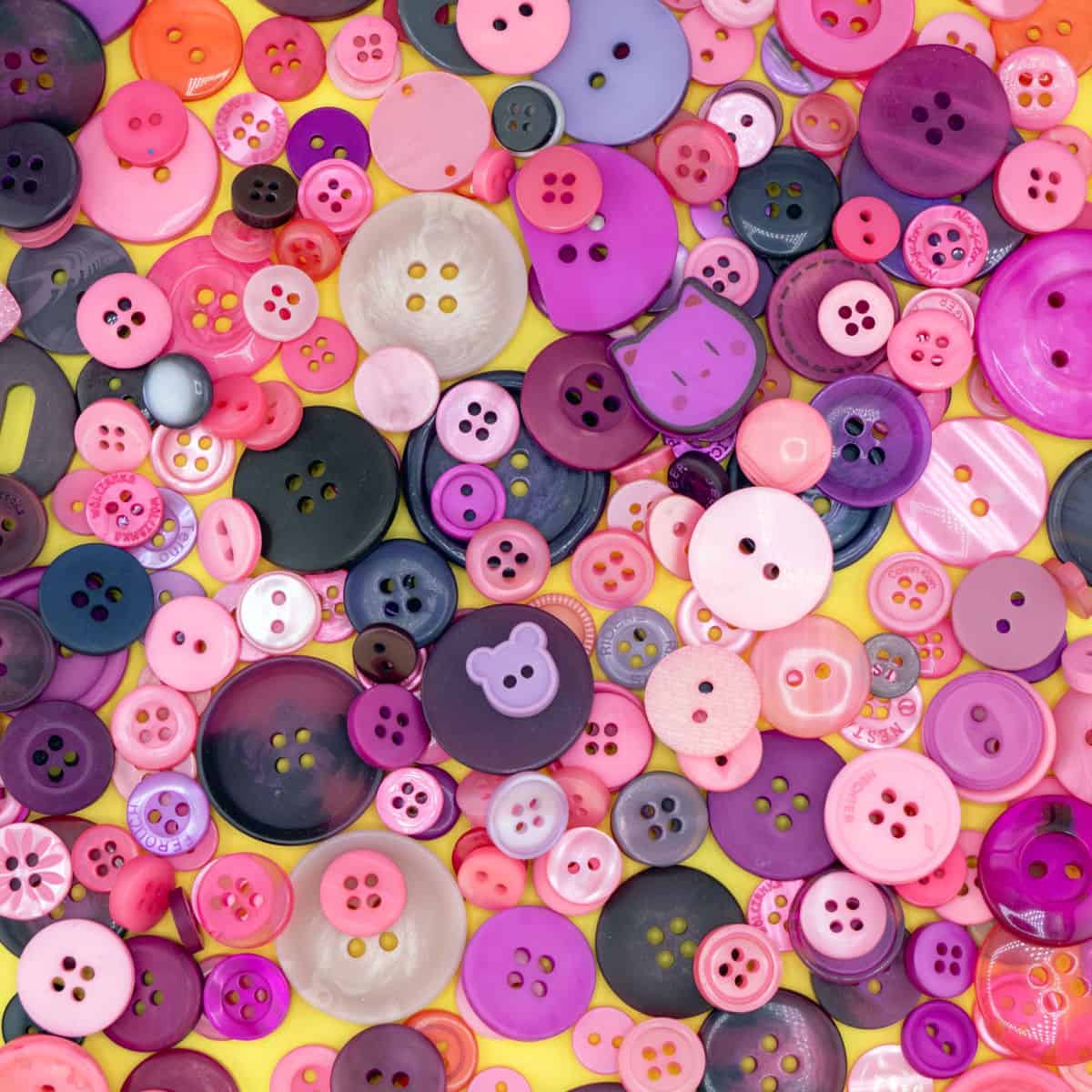 A colorful assortment of pink, purple, and black buttons in various sizes and shapes scattered on a yellow background.