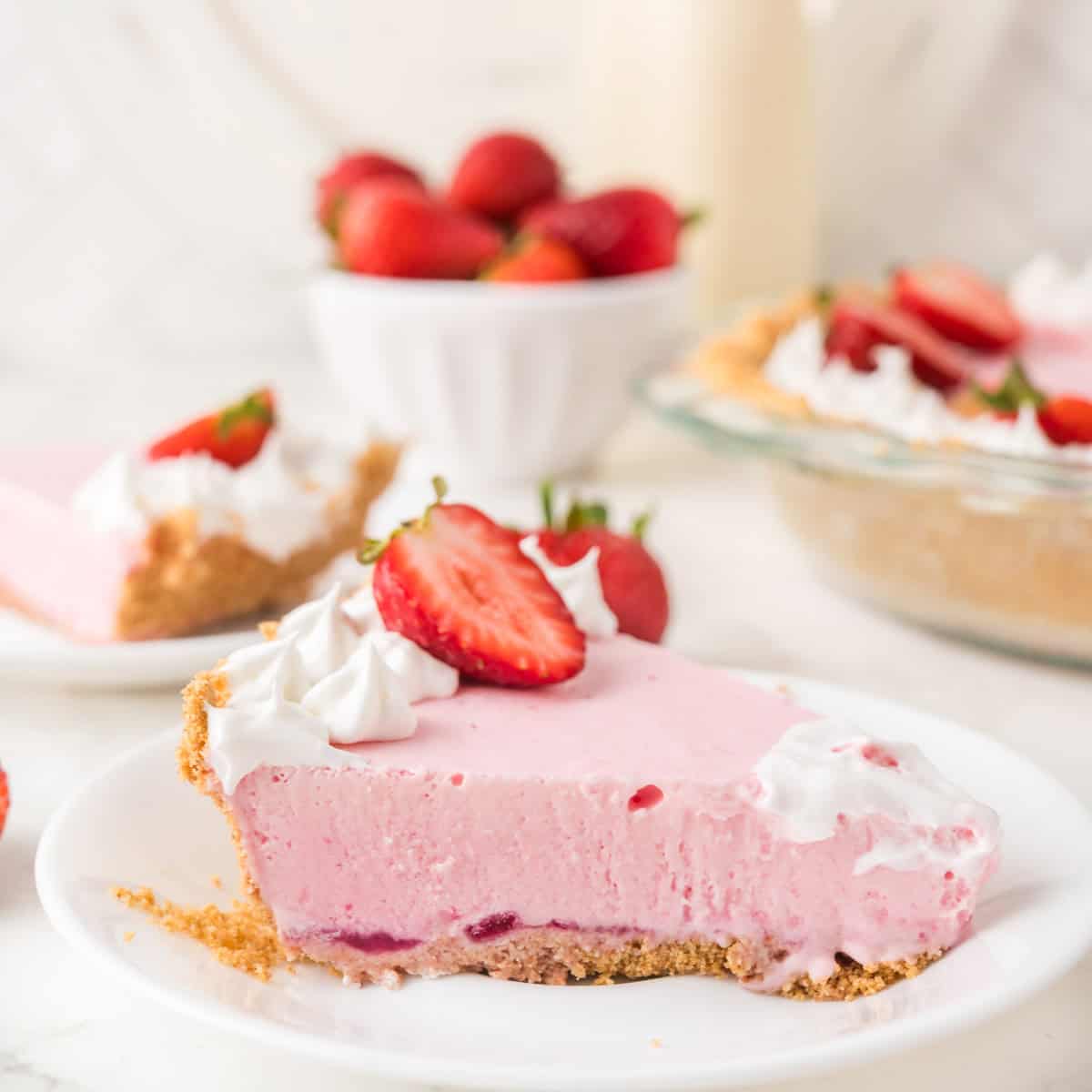 A slice of pink strawberry cream pie with whipped topping and fresh strawberries on a white plate; whole pie and a bowl of strawberries in the background.