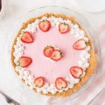 A pink strawberry pie with a graham cracker crust, decorated with whipped cream and fresh strawberry halves, sits on a glass plate on a white table.