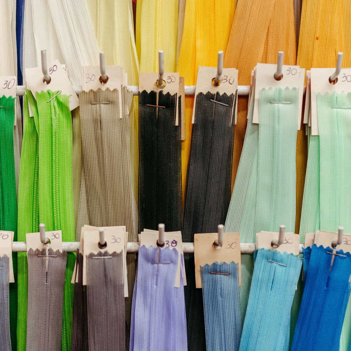 Rows of colorful zippers in various shades, including green, gray, yellow, blue, and black, are neatly displayed on hooks, each bunch labeled with a handwritten “30” on small cards.